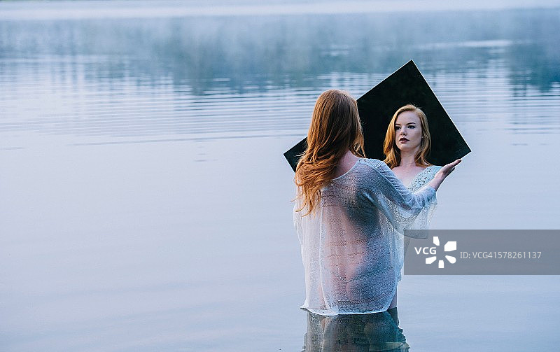 年轻女子站在湖中，手持镜子看着倒影的背影图片素材