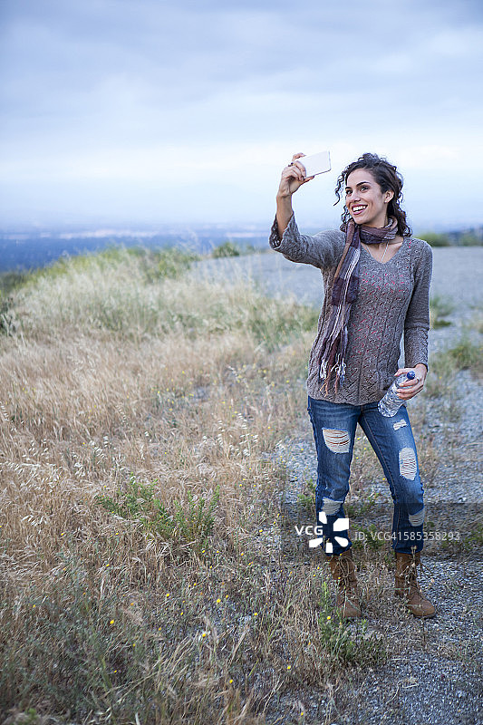在乡村山丘上用智能手机自拍的年轻女人图片素材