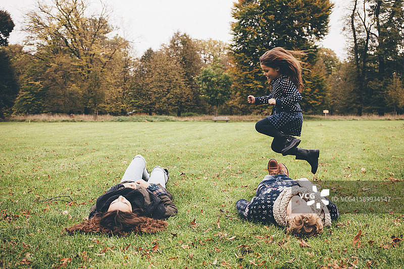 年轻父母躺在草地上，小女孩在公园里跳过他们图片素材