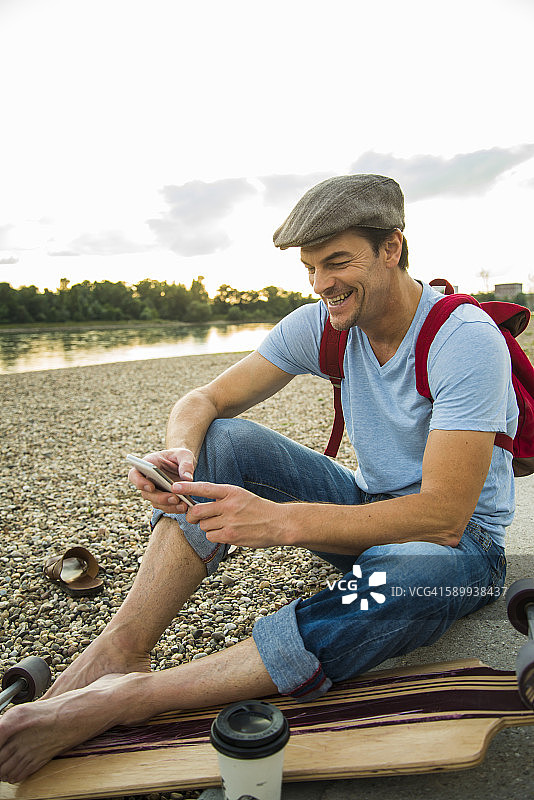 微笑的男人傍晚坐在石滩上，拿着滑板和智能手机图片素材