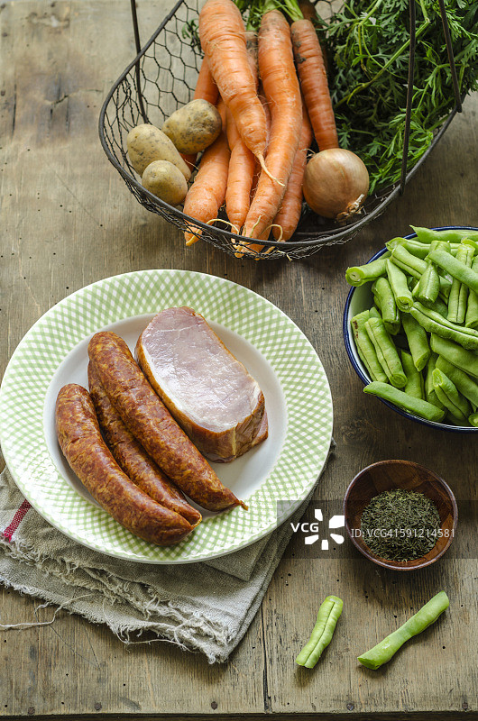 豆 stew 的配料，盘子里有猪肉末香肠和熏猪排，篮子里有胡萝卜、土豆、洋葱和香草图片素材
