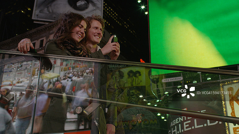 情侣在阳台上拍摄城市十字路口标志性广告牌的仰视角度图片素材