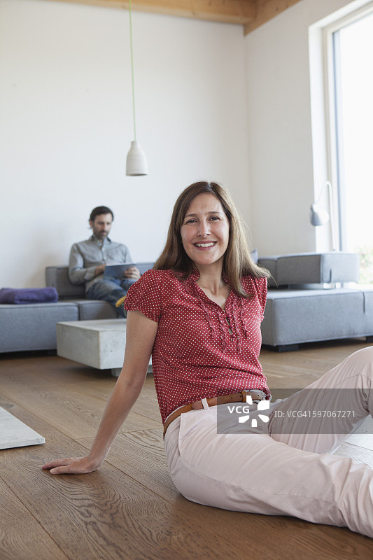 坐在客厅地板上微笑的中年妇女，背景是使用平板电脑的男人图片素材