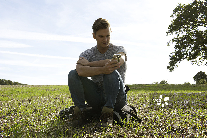 坐在田野背包上阅读智能手机短信的年轻男人图片素材