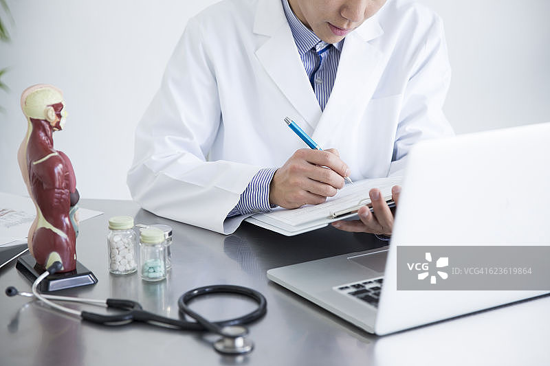 医生一边看笔记本电脑一边书写医疗记录图片素材