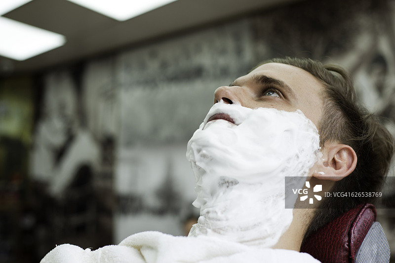 在理发店里，脸上涂满剃须膏向上看的青年男人图片素材