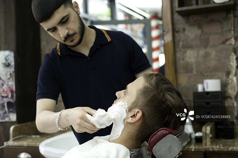 理发店内，年轻男人为顾客涂抹剃须膏图片素材