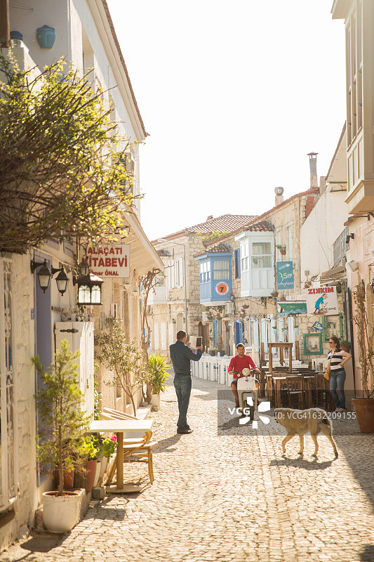在土耳其阿拉恰特，男子使用平板电脑拍摄在鹅卵石街道上骑滑板车的男子图片素材
