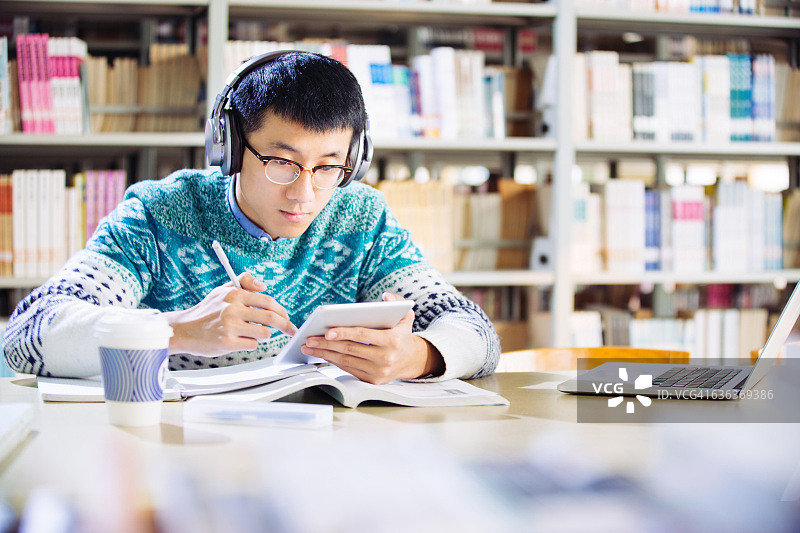 戴着耳机的年轻亚洲男人在图书馆里拿着平板电脑图片素材