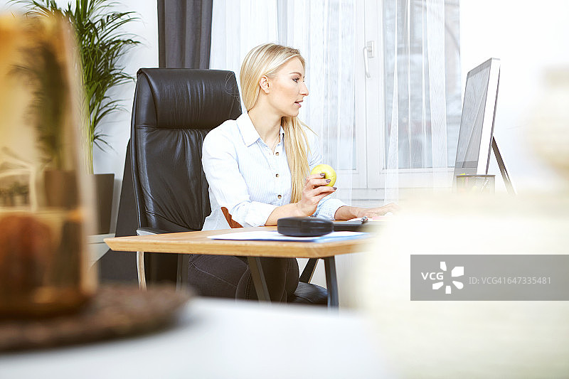 在家里的办公室里坐在桌旁吃苹果的金发女人图片素材