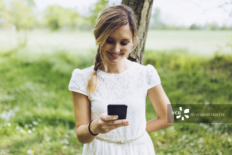 倚靠树干看智能手机的微笑女人图片素材