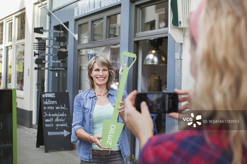 朋友在商店前为拿着“营业中”标牌的女人拍照图片素材