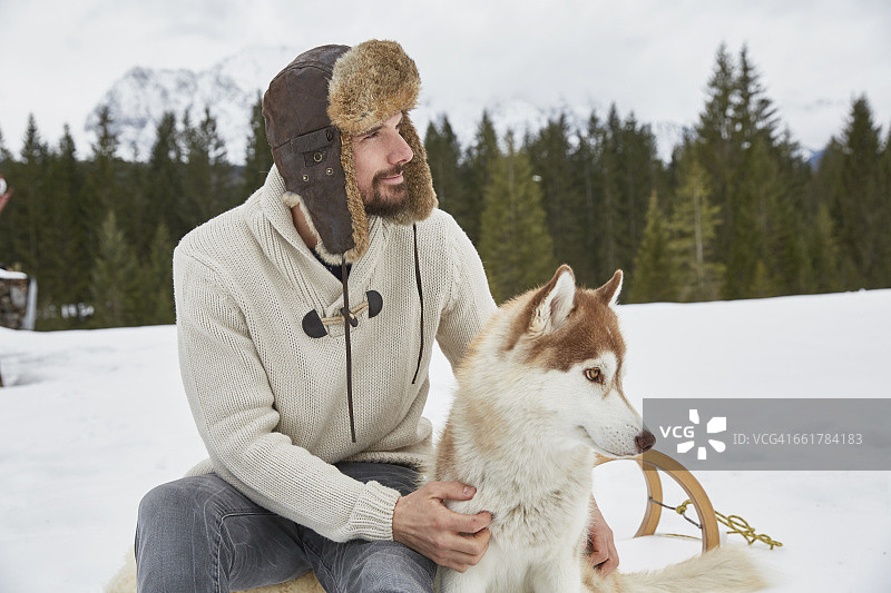 年轻男子戴着飞行帽在德国巴伐利亚埃尔毛的雪地里抚摸哈士奇图片素材