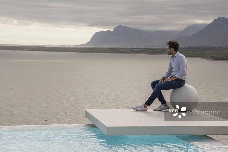 男子坐在湖岸边的石球上图片素材