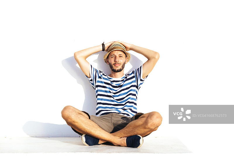 戴草帽穿条纹T恤的度假者靠白墙而坐图片素材
