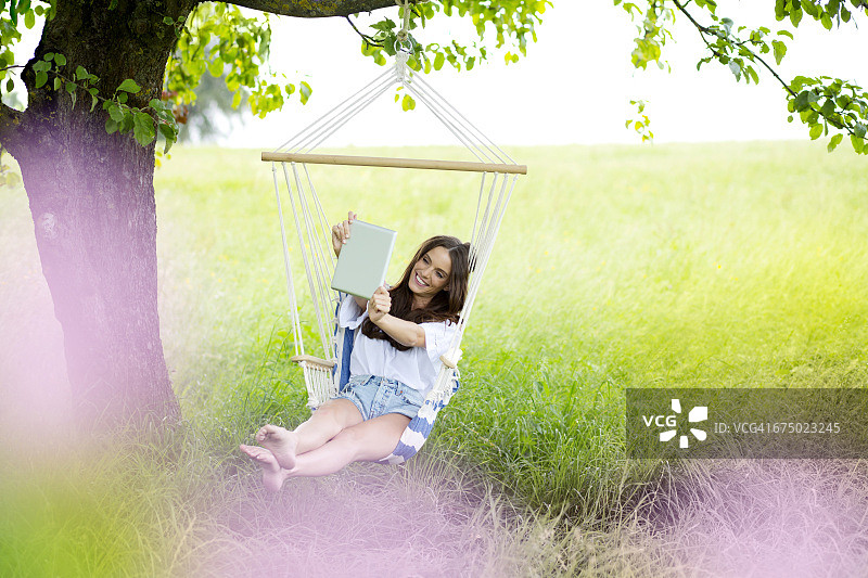 快乐的女人在树下的吊椅上用平板电脑自拍图片素材