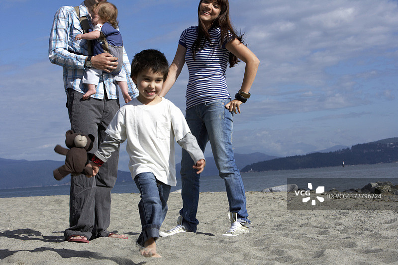抱着泰迪熊的男孩（3-4岁），背景是海滩上带着婴儿（6-9个月）的父母图片素材