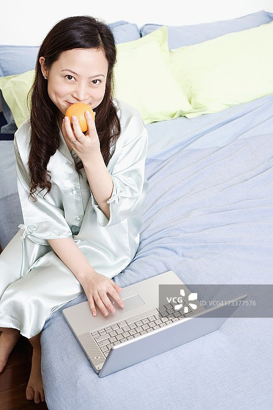 年轻女人在床上用笔记本电脑并拿着橙子的肖像图片素材