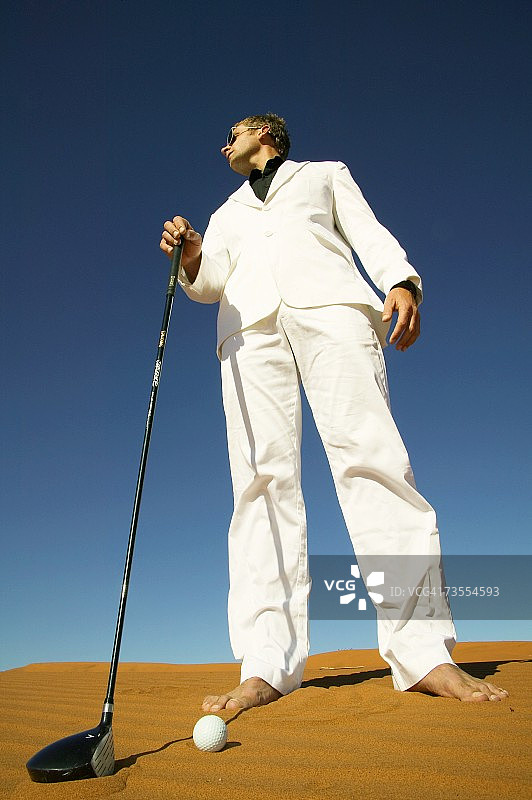在贫瘠沙漠沙丘上，穿着白色西装，手持高尔夫球杆的男人图片素材
