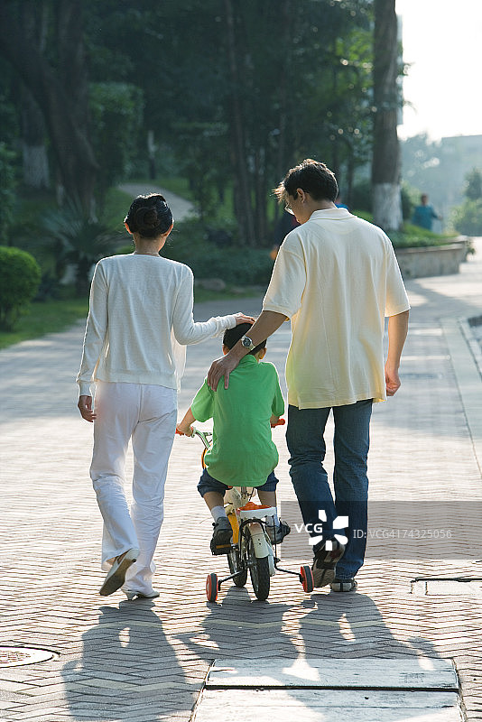 父母陪伴儿子骑带辅助轮的自行车（后视图）图片素材
