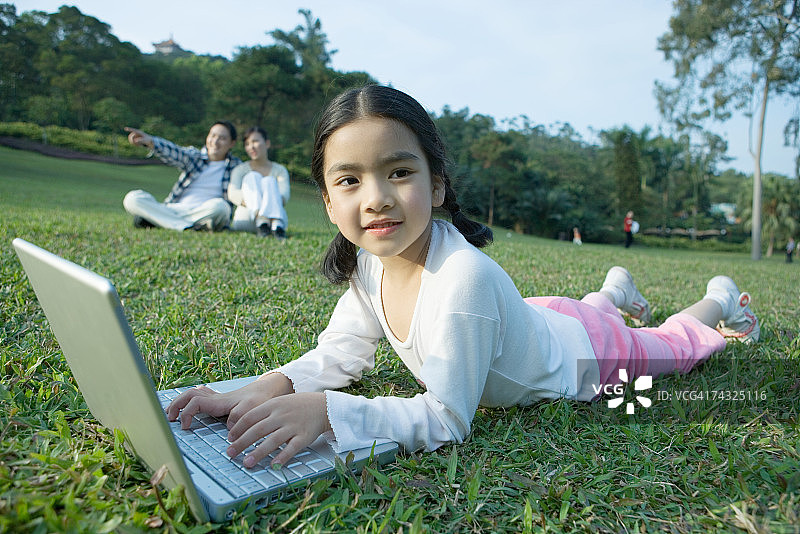 女孩躺在草地上使用笔记本电脑，父母在背景中图片素材
