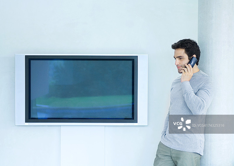 倚靠墙壁，在宽屏电视旁使用手机的男人图片素材