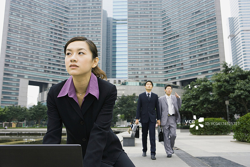 在办公园区使用笔记本电脑的女商人，背景中有两位商人图片素材
