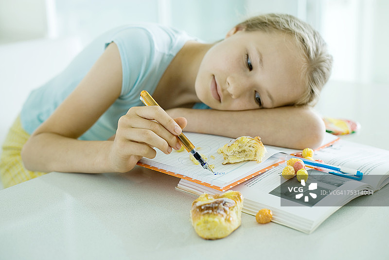 女孩坐在桌旁做作业，头靠在胳膊上涂鸦，食物散落在书上图片素材