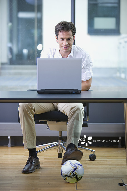 办公室工作人员使用笔记本电脑，桌下放着足球图片素材