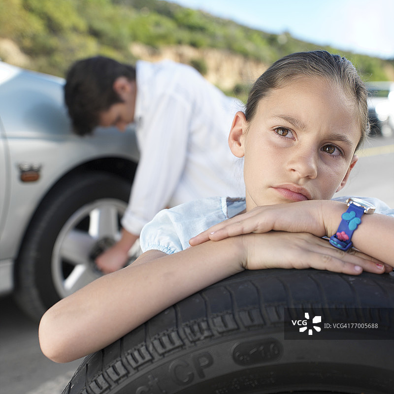 8-9岁女孩拿着备用轮胎，父亲在背景中更换轮胎图片素材