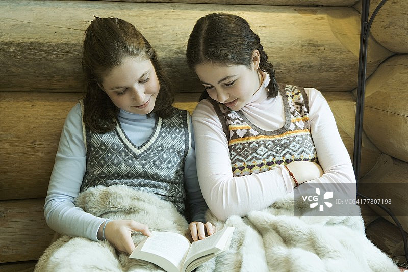 两个穿着冬装的少女一起坐在温暖的毯子下阅读图片素材