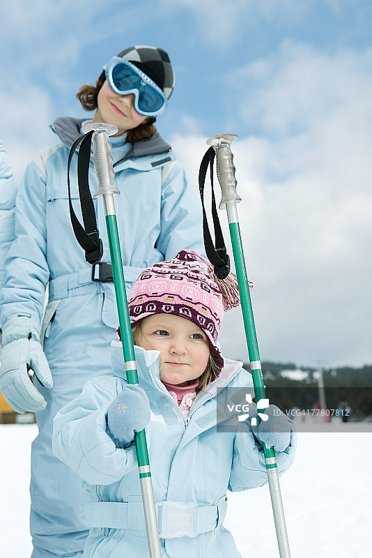 幼儿女孩手持滑雪杖，姐姐站在身后，都穿着滑雪服图片素材