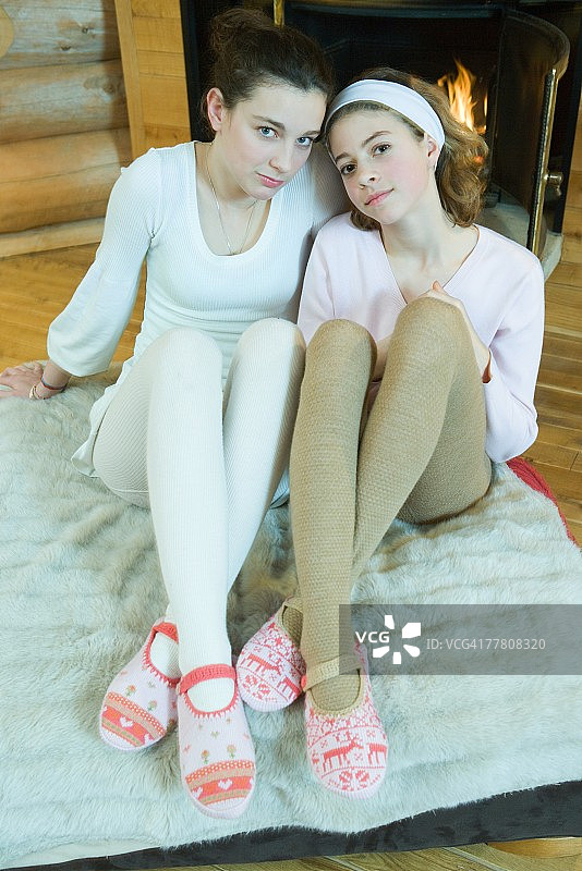 两名少女坐在壁炉旁，穿着紧身裤和拖鞋，看着镜头，全身照图片素材