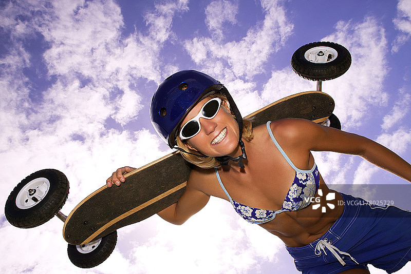 一位扛着山地滑板的女人，倾身看着镜头微笑的低角度照片图片素材
