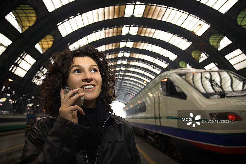 在意大利米兰中央火车站，女子对着手机微笑交谈，背景是驶来的火车图片素材