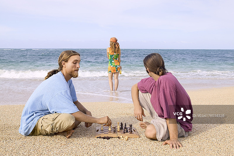两个年轻男人在海滩上下象棋；一个在走棋，另一个在看站在水边的一位年轻女士图片素材
