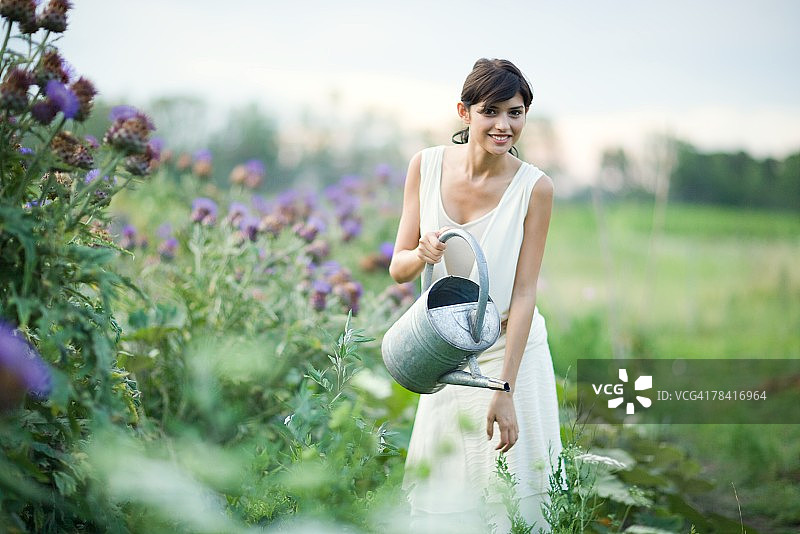 年轻女人在花园里用水壶浇花，对着镜头微笑图片素材