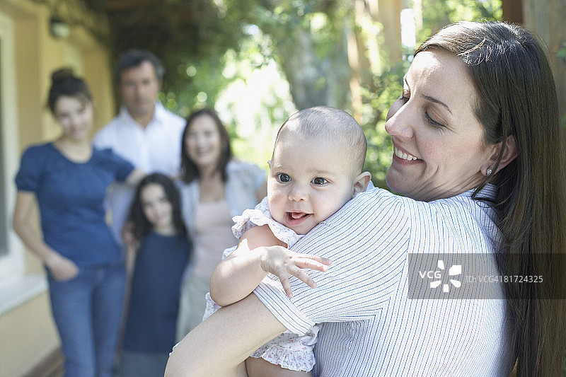 户外抱着宝宝微笑的女人，背景是家人图片素材