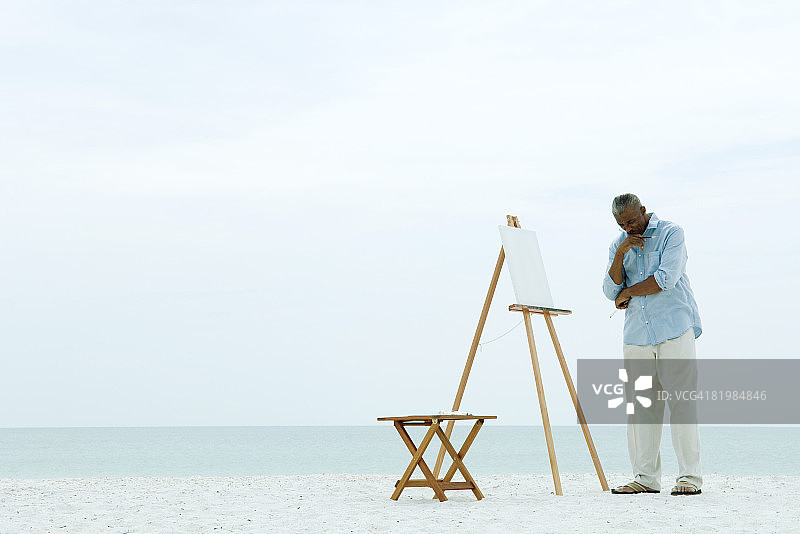 站在海滩空白画布前，手托下巴向下看的男性画家图片素材
