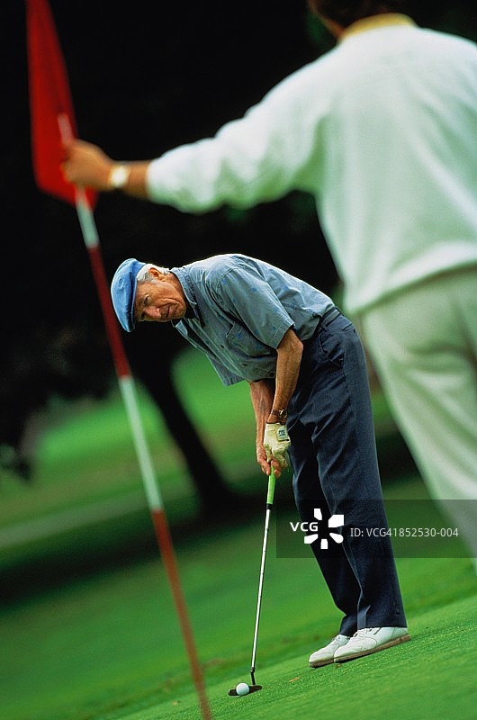准备推杆的老年高尔夫球手，球洞前方有人拿着旗子图片素材