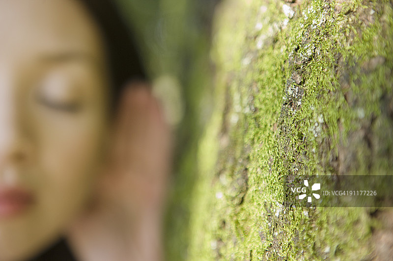 靠着树，手放在耳朵上，闭着眼睛，前景聚焦的女人图片素材