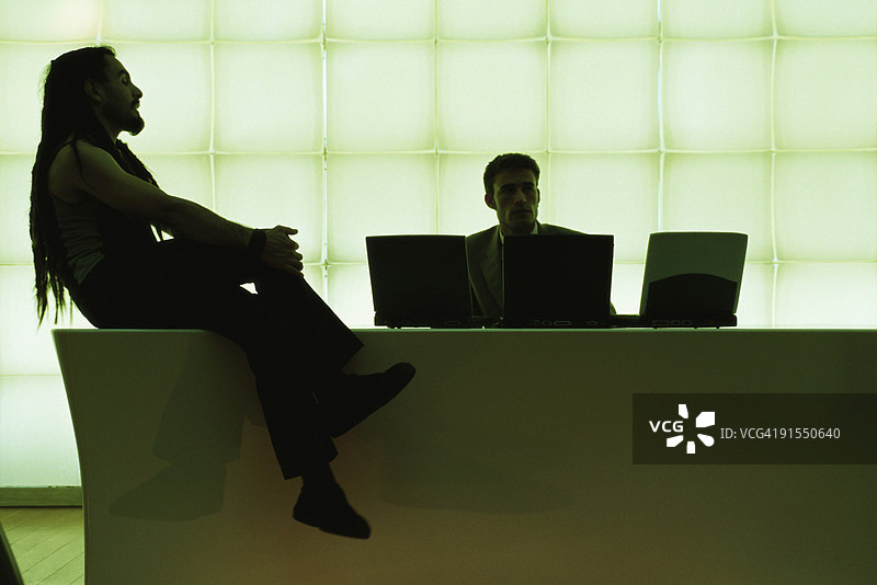 留着脏辫的男子双腿交叉坐在桌旁，与使用笔记本电脑的商人交谈图片素材