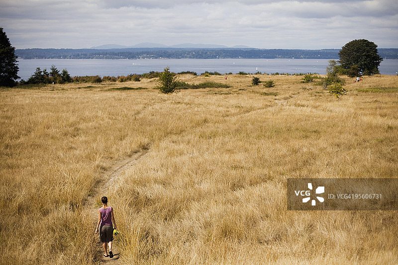 年轻女子身穿紫色衬衫，带着绿色瑜伽垫穿过金色草地图片素材