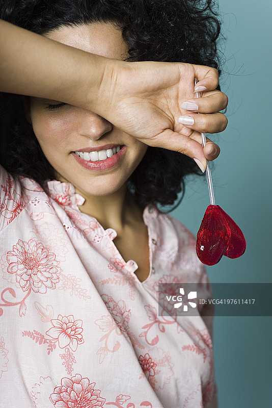 面带微笑，用手臂遮住眼睛，拿着心形棒棒糖的女人图片素材