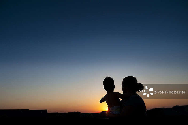 黄昏时分，阿姨抱着指向郊区日落的幼儿的剪影图片素材