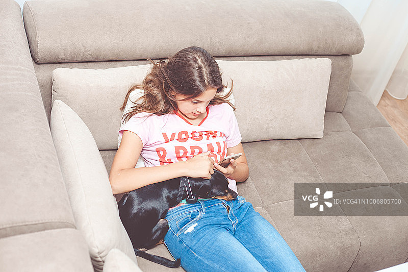 在沙发上抱着宠物狗看智能手机的少女图片素材