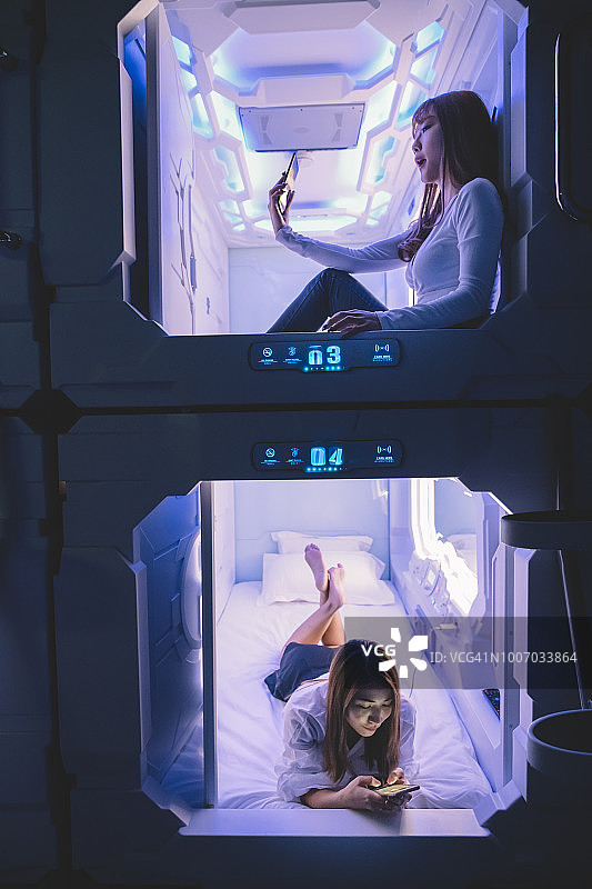 两位中国亚洲女游客在胶囊酒店房间办理入住，用手机上网和自拍图片素材