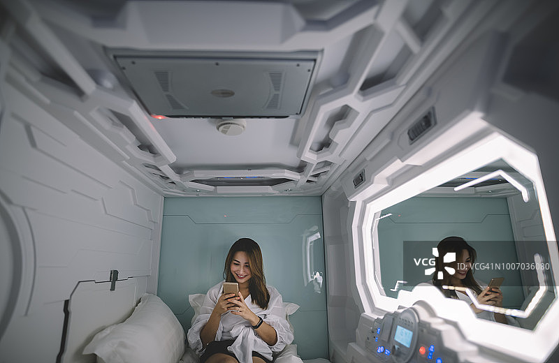一位亚洲中国女游客在太空舱酒店房间办理入住手续，并在床上用手机上网图片素材