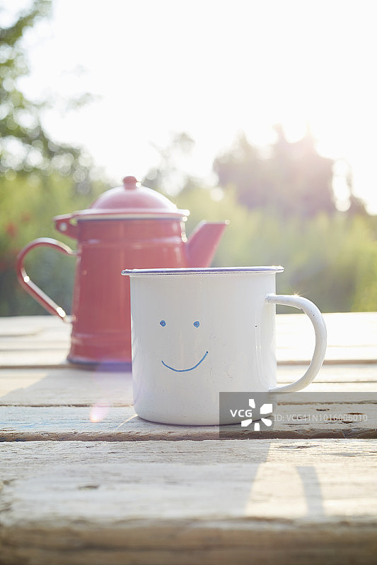 花园里木桌上，带有笑脸图案的搪瓷杯和咖啡壶特写图片素材