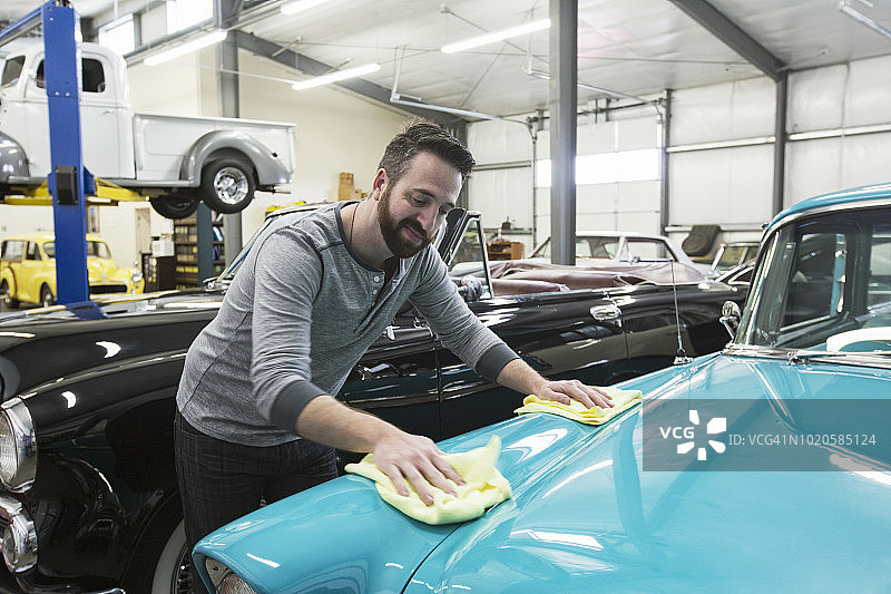 一位白人男汽车修理工在他的老爷车修理店里给一辆旧轿车的引擎盖打蜡图片素材